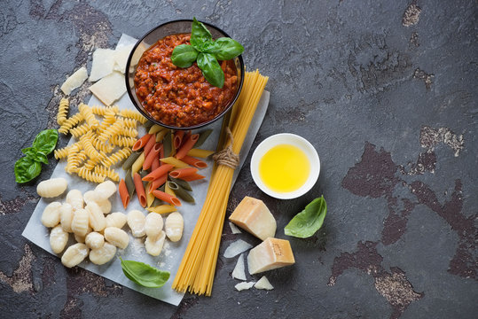 Bolognese sauce with different types of raw pasta and potato gnocchi, high angle view with space on a brown stone background