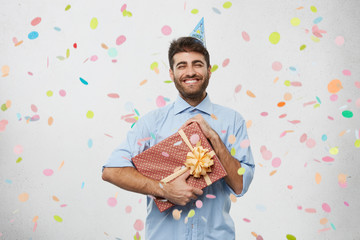 Portrait of cheerful joyful young Caucasian man with beard enjoying his birthday party, standing isolated in light room with confetti falling down on him, smiling, holding present in beautiful paper