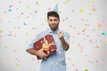 Portrait of greedy young European unshaven male grimacing, holding tight gift in wrapping paper, making fig sign at camera as he doesn't want to show birthday presents to his guests. Body language