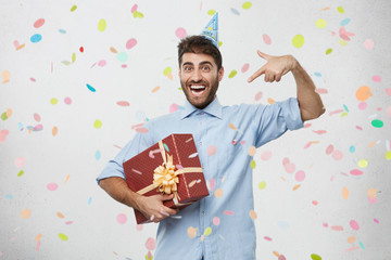 Ecstatic overjoyed young Caucasian man with holiday cap on head holding large box in wrapping...