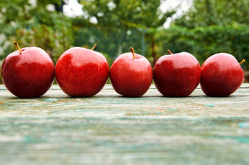Five red apples on wooden green brown aged texture surface close up. Apples on blurred nature background. Side view