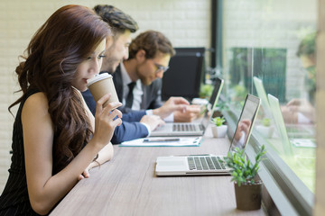 Businesswoman drinking coffee break looking at her laptop to work while coworker interacting in the background in the office