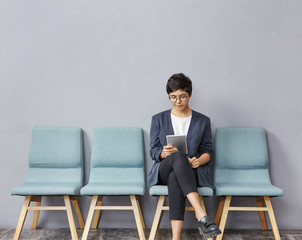 Confident successful young businesswoman with short hairstyle wearing stylish clothes and glasses using digital tablet while waiting for doctor's appointment, sitting on chair in hall at clinic - obrazy, fototapety, plakaty