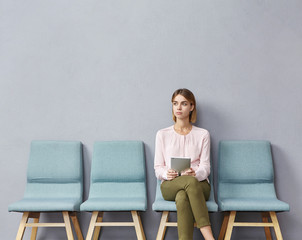 Portrait of confident serious young European female sitting on chair in waiting room with electronic tablet, setting her mind up before job interview or meeting with potential business partners - obrazy, fototapety, plakaty