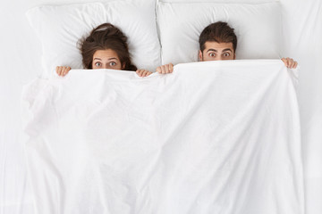 High angle indoor shot of funny adult couple lying in bed with blanket pulled up over their noses...