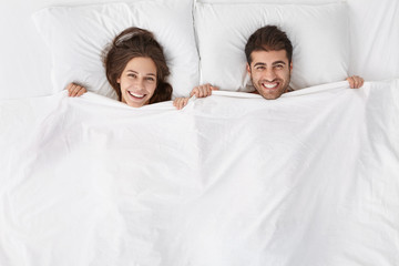 Top view of heads of happy lovely couple lying in bed and smiling broadly after good sex. Lovers enjoying intimacy and togetherness. Just married spending first night at hotel room, feeling excited