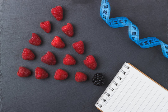 Red raspberries, tape measure, notebook and a pen on the black slate stone background