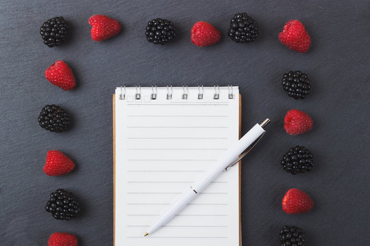Red raspberries, tape measure, notebook and a pen on the black slate stone background