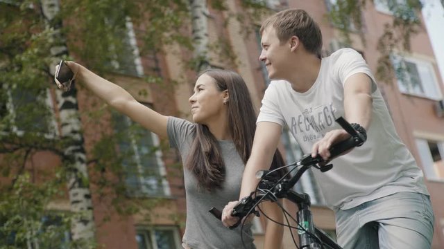 Young couple in love walking in a city park with bicycles and taking pictures. A guy and a girl take pictures of themselves on a smartphone, making selfies. Birches and houses on a background.