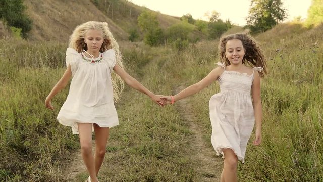 Close up view of happy children in a white dresses dancing, whirling and laughing. Close up view of two little girls whirling in a round dance in a bright sunset. Cute smiles. Childhood games.