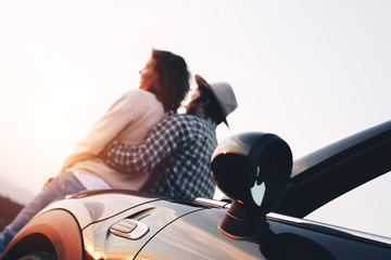 Trendy modern loving couple sits on hood of cabriolet and hugs, looks like sunset
