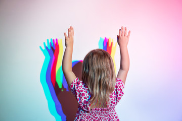 Little girl stands against the wall with her arms raised with her back to the camera. On the wall is a diffraction shadow - obrazy, fototapety, plakaty