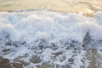Dettaglio della schiuma delle onde del mare quando si infrangono sulla costa. La spaiggia è dorata e sabbiosa; il mare è blu e agitato. Il sole è quasi tramontato e si specchia nell'acqua.