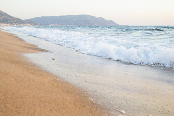 Dettaglio di un tratto di costa sabbiosa con in fondo un promontorio. Il mare è mosso e agitato e le onde si infrangono sulla spiaggia sabbiosa con violenza creando la schiuma.