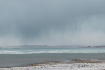 panoramic view of watery surface of lake with snow on coast and storm clouds in sky
