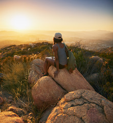 Rear view of woman hiker sitting on rock on top of hill while looking at sunset over San Diego...