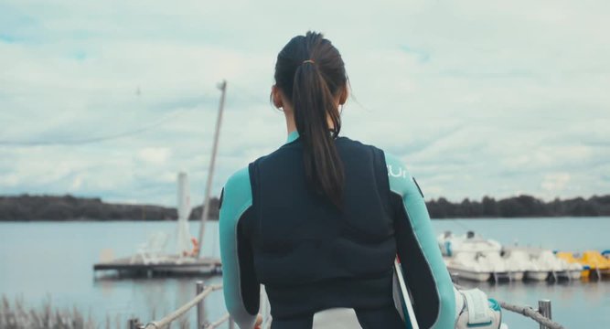 TRACKING back view young female in a swimsuit walking towards water with a wakeboard in hands, cloudy sky. 4K UHD 60 FPS 