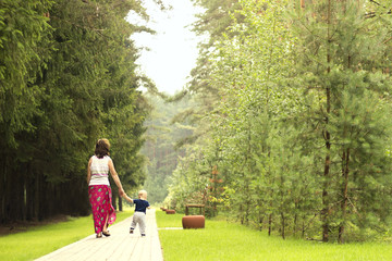 Fototapeta premium Mom and son on the walk in the park. Back view of walking family. First independent steps of one year toddler.