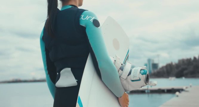 TRACKING back view young female in a swimsuit walking towards water with a wakeboard in hands, cloudy sky. 4K UHD 60 FPS 