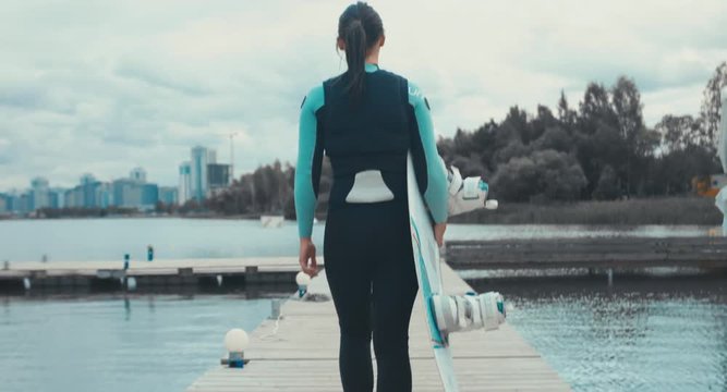 TRACKING back view young female in a swimsuit walking towards water with a wakeboard in hands, cloudy sky. 4K UHD 60 FPS 