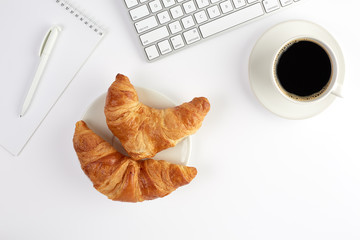 Top view of keyboard, blank notebook with pen, two croissants and cup of coffee on white background