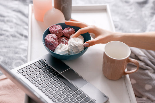Unrecognizable woman takes zephyr from teal plate. Early breakfast with cup of tea and sweets combining with chatting on laptop, close up