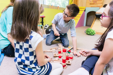 Cute pre-school boy cooperating with his colleagues at the construction of a structure made of...