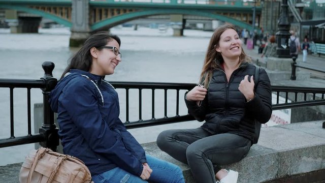 Two girls sit at the banks of River Thames and have small talk
