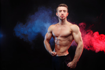 Bodybuilder man with perfect abs, shoulders,biceps, triceps and chest flexing his muscles in blue and red smoke. Strong young athlete with six pack - obrazy, fototapety, plakaty