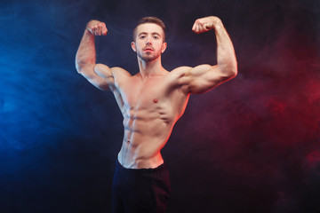 Strong young man bodybuilder demonstrating huge muscles on smoke background. Man with perfect abs,...