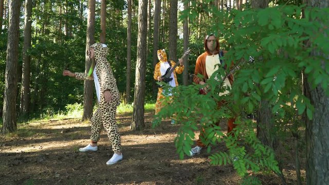 Mad people in animals costumes dances, jumps, have fun in forest on grass. Guys and girl musicians dressed the costumes of zebra, jaguar, giraffe, bear, squirrel plays music with guitar, bass, drums