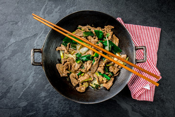 Traditional chinese mongolian beef stir fry in chinese cast iron wok with cooking chopsticks, stone slate background. Top view, copy space