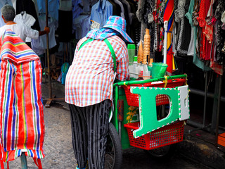 Thai local traditional street food ice cream stall cart beside clothing shop market on paved road, seller man bending scooping in sunny weather