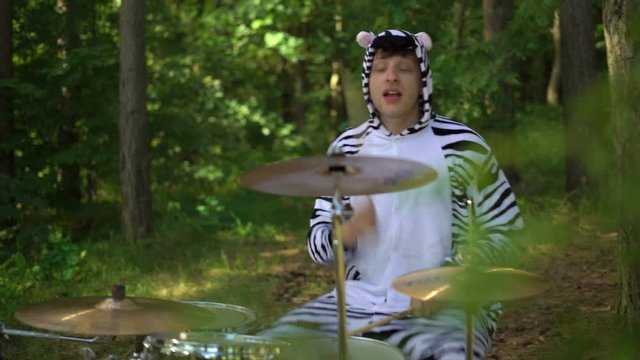 Man in zebra costume plays on drums, smiles, enjoys, go crazy in forest between trees. Drummer is playing music on the grass