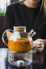 Sea buckthorn tea in the glass teapot.The young blonde girl drinking tea in the cafe on the background