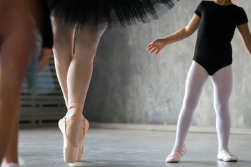 Close-up of an adult beautiful ballerina in a black dress, white pantyhose and pointe shoes is dancing, and a little girl of a ballerina who is just beginning to study at her
