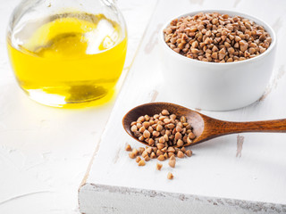 Brown buckwheat in spoon and buckwheat oil in glass bottle on white wooden background. Copy space.