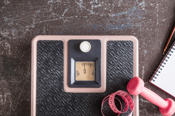Old weight scale, dumbbell, measuring tape and notebook on the dark floor in the gym. Cares about a...