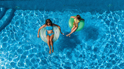 Aerial top view of children in swimming pool from above, happy kids swim on inflatable ring donuts and have fun in water on family vacation
