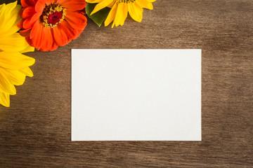 Naklejka na ściany i meble White blank greeting card with yellow and red flowers on the brown wooden table. Empty place for a text. Top view.