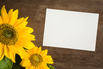 White blank greeting card with beautiful yellow sunflowers on the brown wooden table. Empty place for a text. Top view.