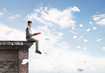 Handsome student guy on roof edge reading book and paper planes fly around