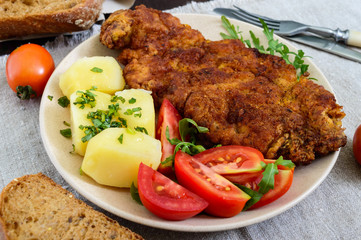 Golden schnitzel, boiled potatoes and salad with tomatoes on a plate on a dark wooden background.