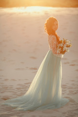 Fototapeta na wymiar Bride with bouquet stands on sand in desert. Backlight and pastel colors.