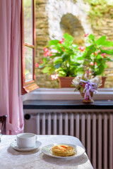Daylight view to pancakes with syrup and a white cup on table. Window and flower background.