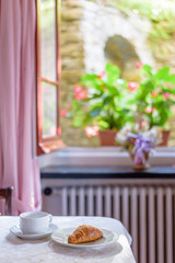 Daylight view to fresh croissant and a white cup on table. Window and flower background.