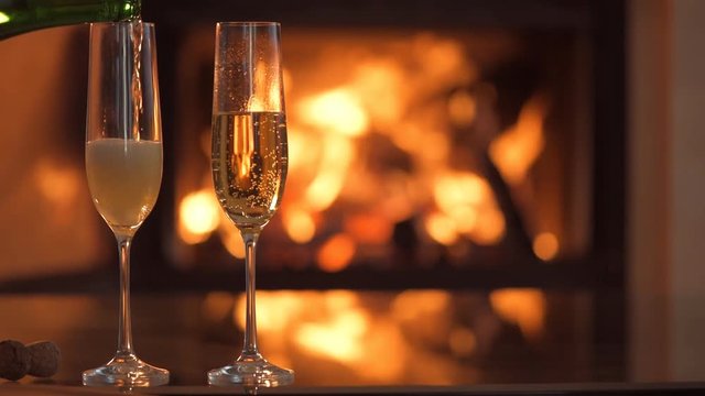 Champagne in two glasses on table in front of burning fireplace.