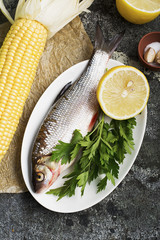 The raw ingredients are fish and vegetables for cooking. River fish of carp breeds with onion, lemon, greens, corn, zucchini, salt, olive oil, basil for recipe. On a gray background. Top View.