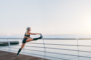 Young beautiful athletic girl with long blond hair in headphones listening to music and doing stretching at sunrise over the sea