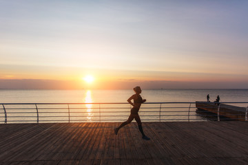 Fototapeta premium Silhouette of a young beautiful athletic girl with long blond hair in headphones, who listens to music and runs at dawn over the sea